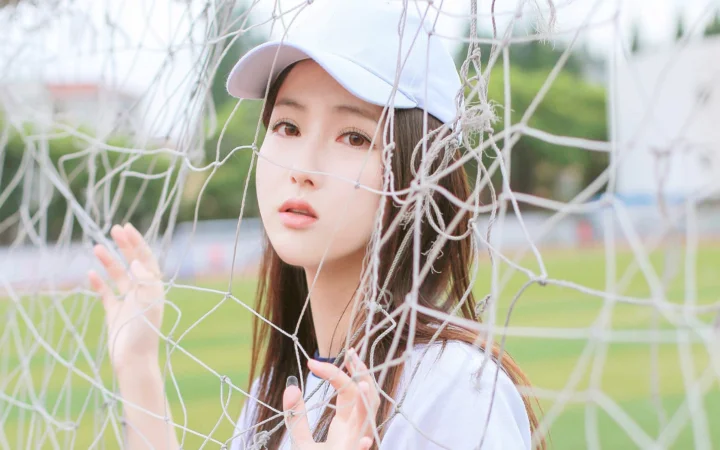 淡い色の帽子をかぶった女性が、緑のフィールドを背景にサッカーゴールのネットの後ろに立っています。