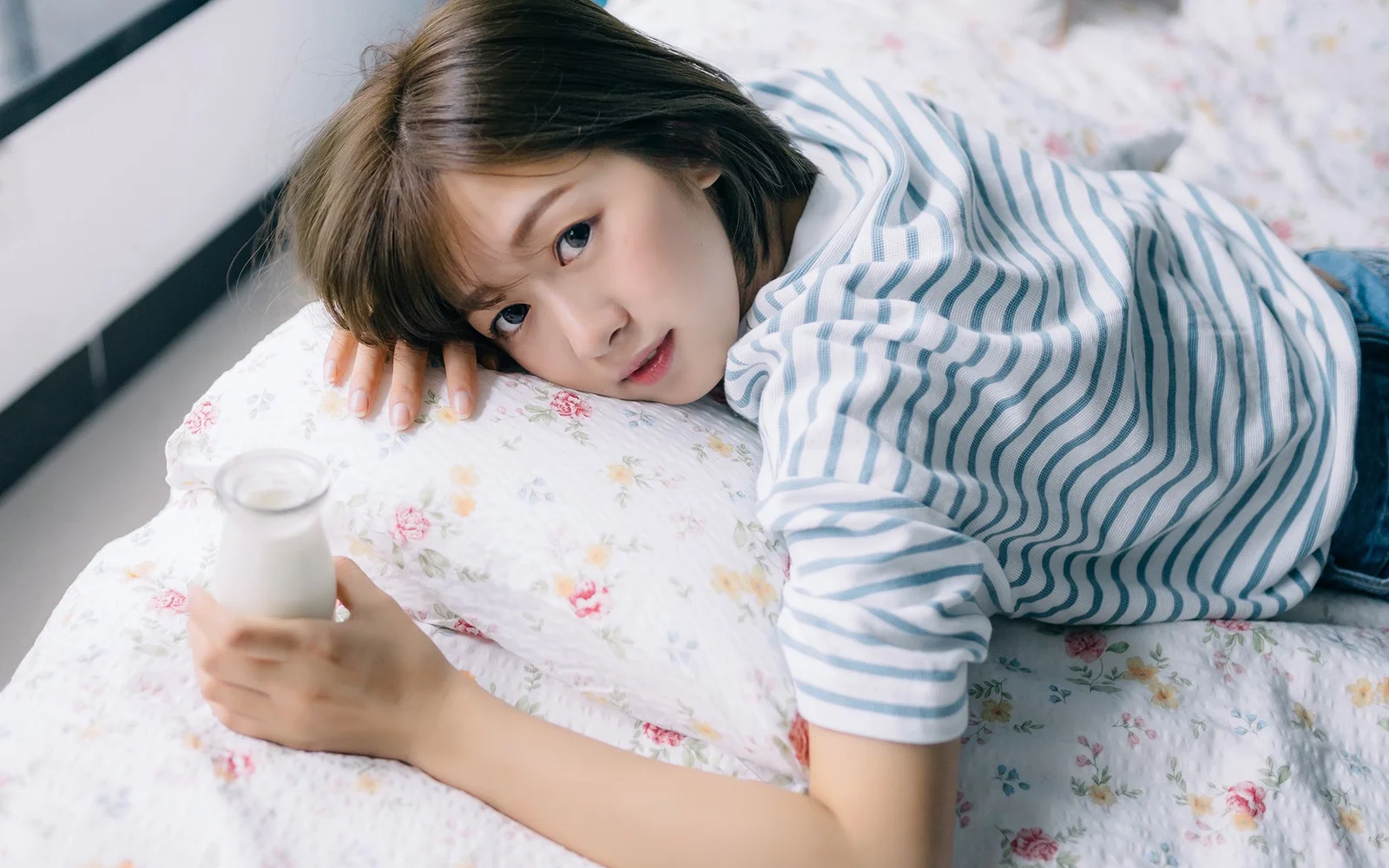 一名成年女性穿着蓝白条纹上衣，趴在铺有花卉图案床品的床上并看向镜头。