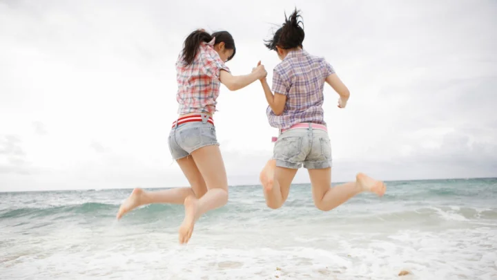 两名女性在海边的浅水中牵手跳起，背景是海面与阴云天空。