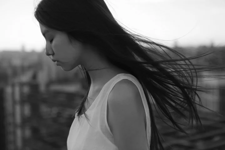 흑백 화면 속 긴 머리의 여성이 흐릿한 도시를 배경으로 옆모습을 보이며 머리카락이 바람에 흩날리고 있다.