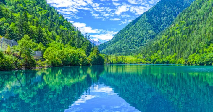碧绿色湖泊倒映着周围的森林与山峦，远处可见蓝天白云。