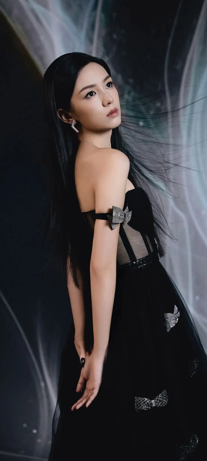 검은 드레스를 입은 여성이 어두운 배경 앞에서 뒤돌아 카메라를 바라보고 있다.
