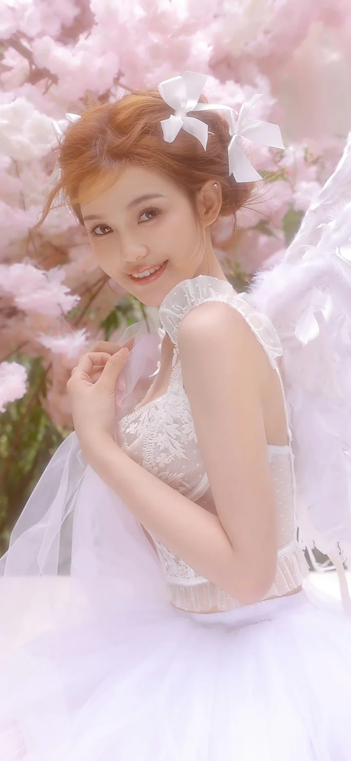 一名身穿白色连衣裙、头戴蝴蝶结的女子站在粉色樱花间微笑回望镜头。