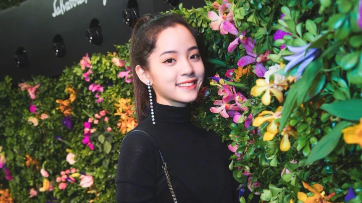 一名身穿黑色高领上衣的女子站在色彩缤纷的花墙旁微笑。