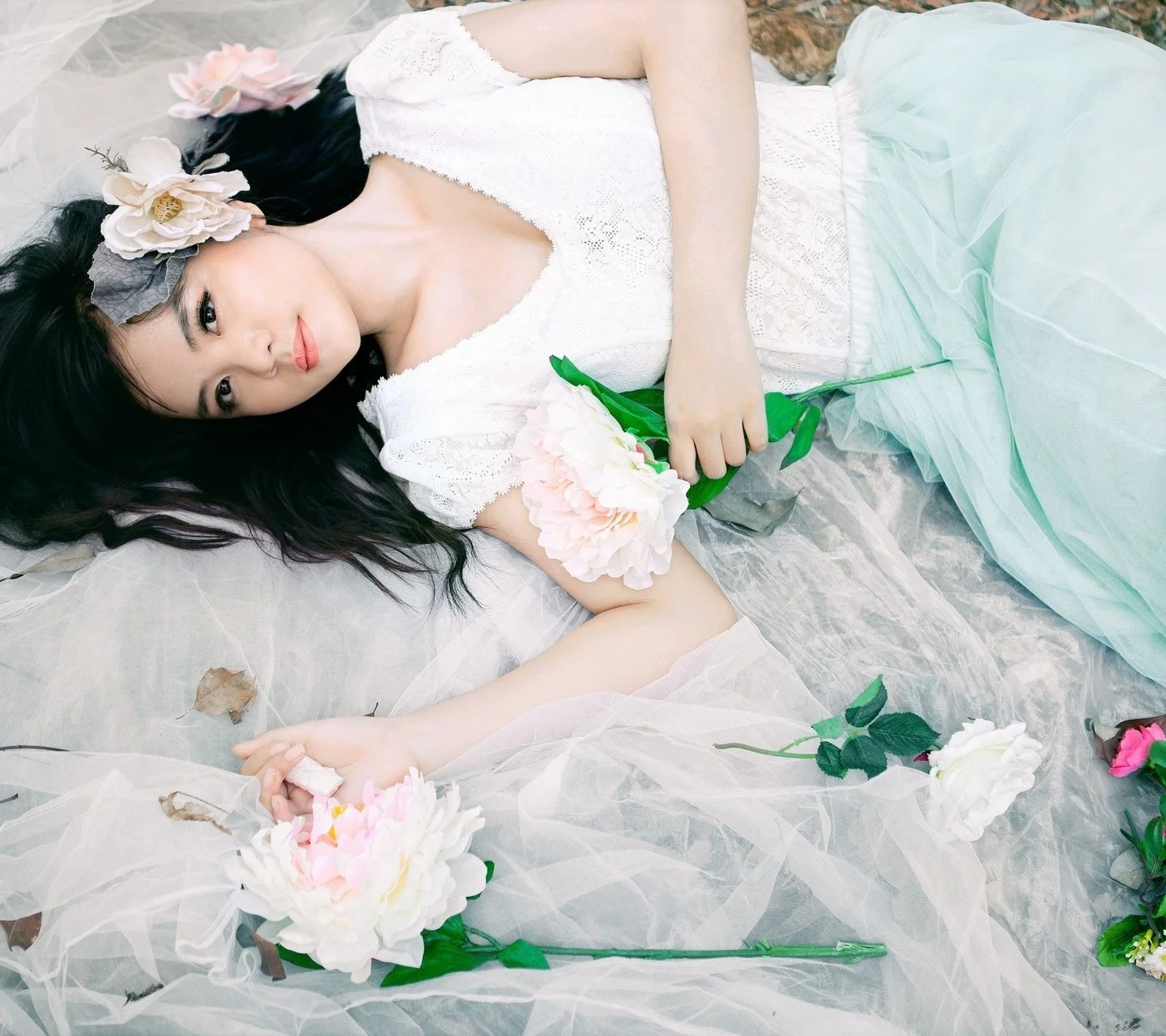 白いドレスを着た女性が、薄い布と花に囲まれて横たわり、花束を手にしている。