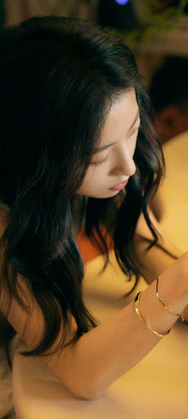 긴 머리의 여성이 따뜻한 빛 속에서 노란색 상의를 입고 아래를 바라보고 있다.