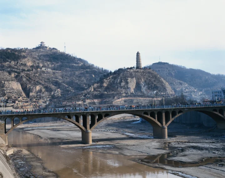 一座拱桥横跨山谷前的河道，远处山坡上可见一座塔和传统建筑。