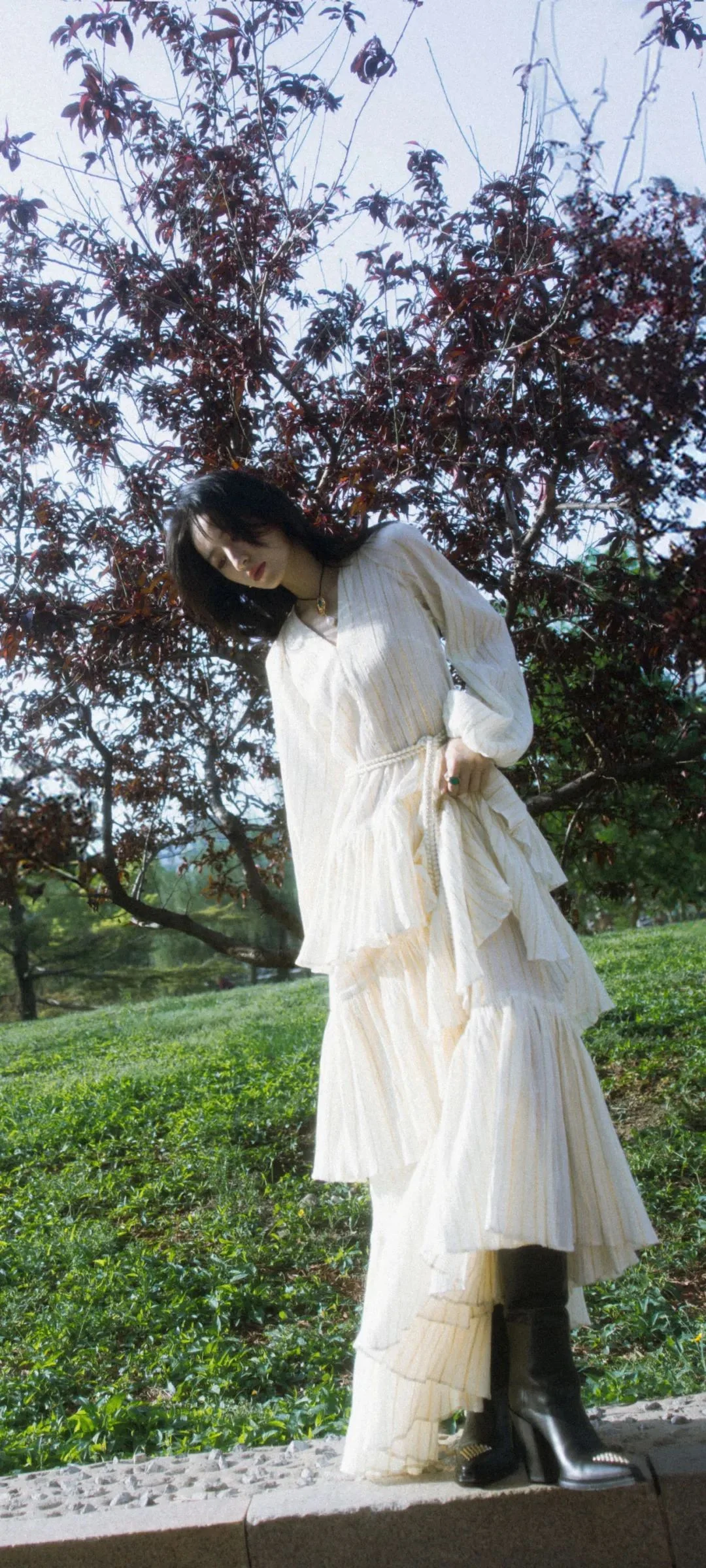 白いロングドレスを着た女性が木々と草地のそばに立っている。