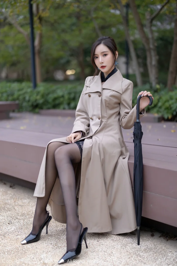 밝은 색 트렌치코트를 입은 여성이 공원 벤치 옆에 앉아 검은 우산을 들고 있습니다.