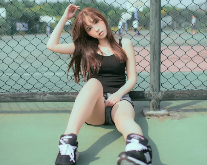 一名女性坐在运动场的铁丝网围栏前，穿着深色上衣、短裤和运动鞋。