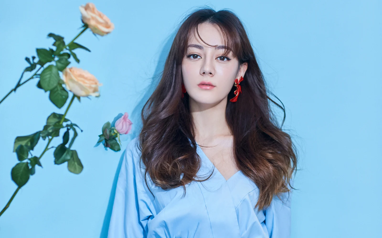 一名长发女性穿着浅蓝色上衣站在蓝色背景前，左侧可见几枝玫瑰。
