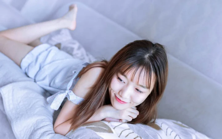 一名棕色长发女子侧躺在床上，微笑着看向镜头。