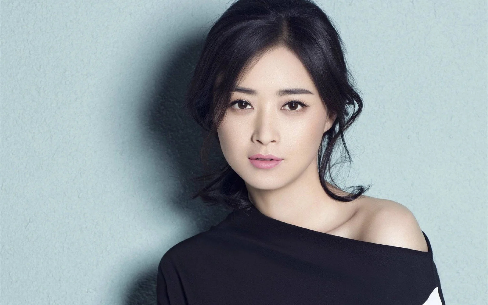 검은 머리의 여성이 검은색 오프숄더 상의를 입고 연한 파란색 배경 앞에서 카메라를 바라보고 있습니다.