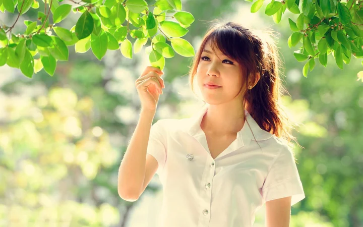 一名成年女子站在绿叶掩映的户外环境中，面带微笑并抬手触碰枝叶。