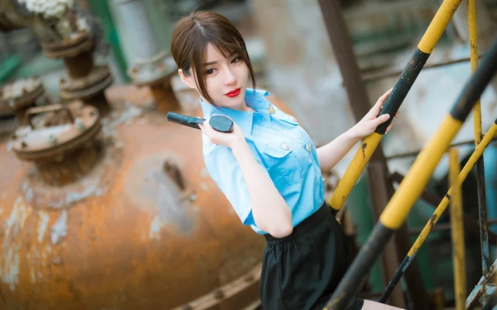 一名身穿浅蓝色上衣的女性站在工业设施旁，手持工具并望向镜头。