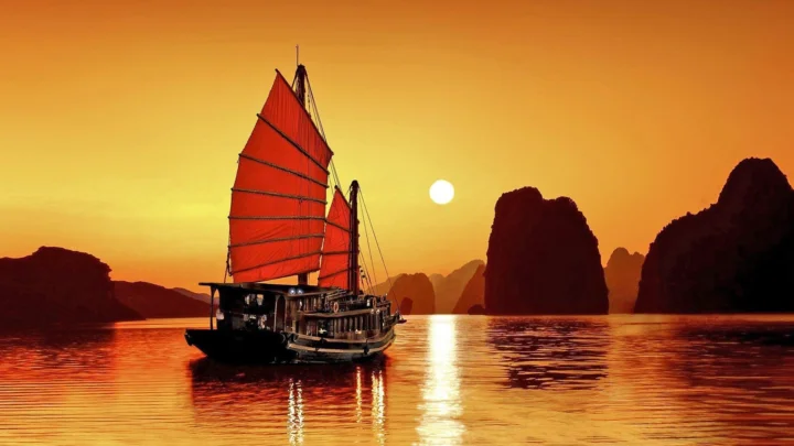 夕阳映照下，一艘红帆木船行驶在群山环抱的水面上。