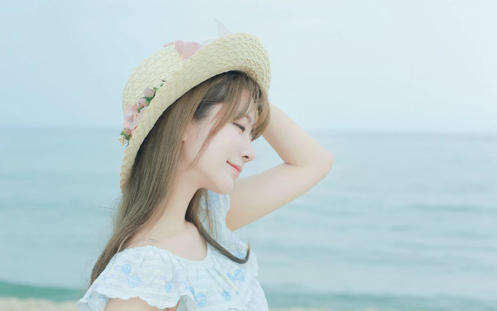 밀짚모자를 쓴 여성이 잔잔한 푸른 바다를 배경으로 해변에 서 있다.