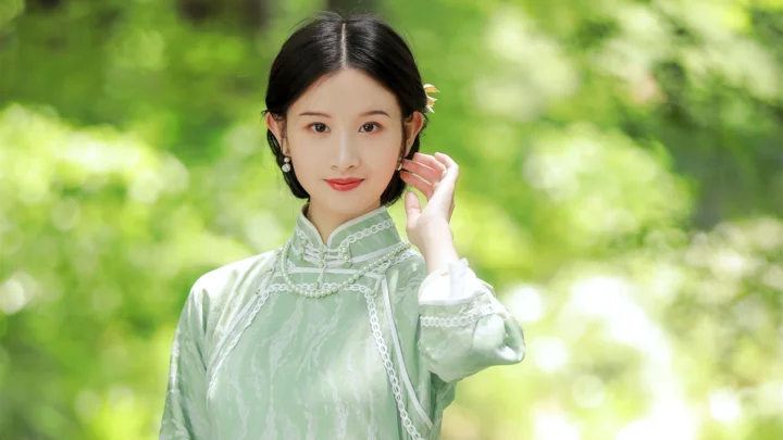 一位身穿浅绿色传统风格服饰的女性站在明亮的绿色背景前，手轻触耳侧。
