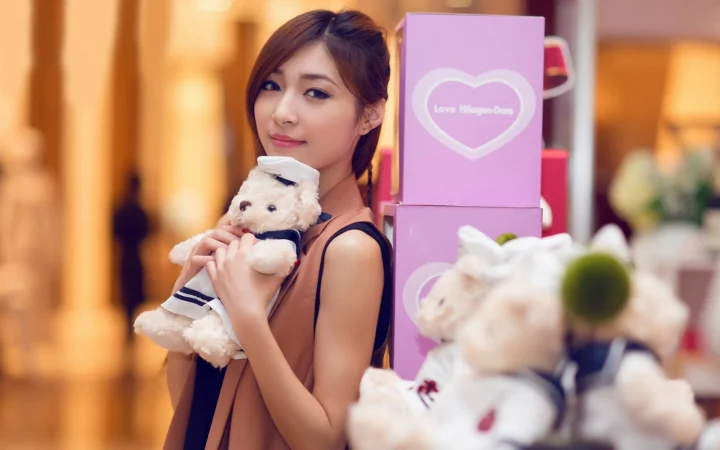 一名女子抱着泰迪熊，身旁摆放着粉色盒子和更多玩偶。