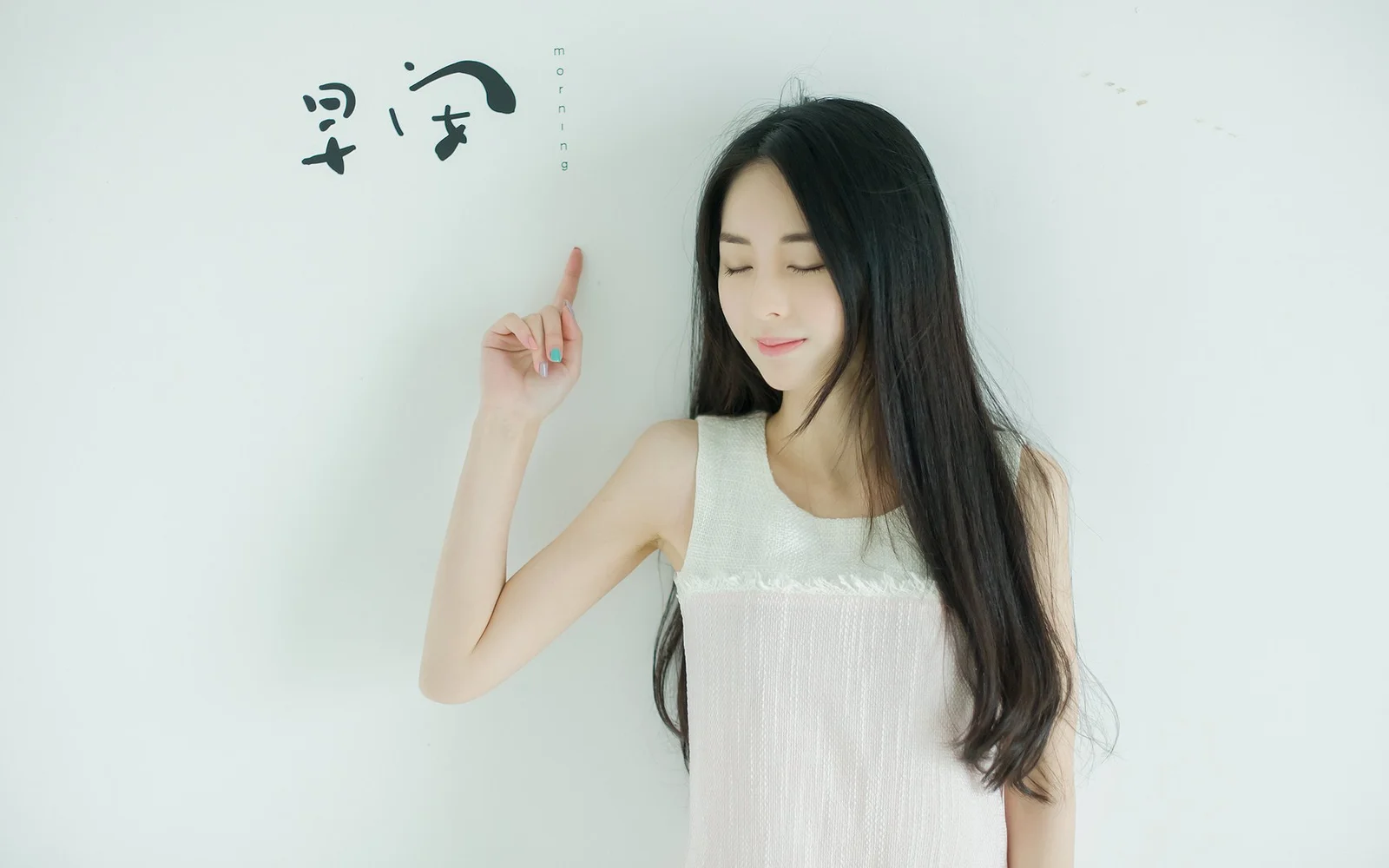 一名身穿白色无袖连衣裙的女子闭着眼睛站在浅色背景前，并抬手指向上方。