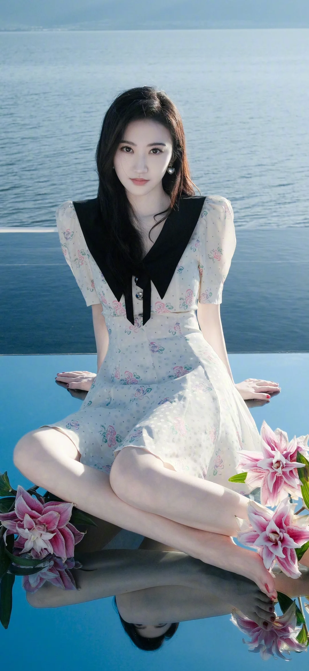 一名成年女性穿着浅色花卉连衣裙坐在水边，周围点缀着鲜花，水面映出她的倒影。