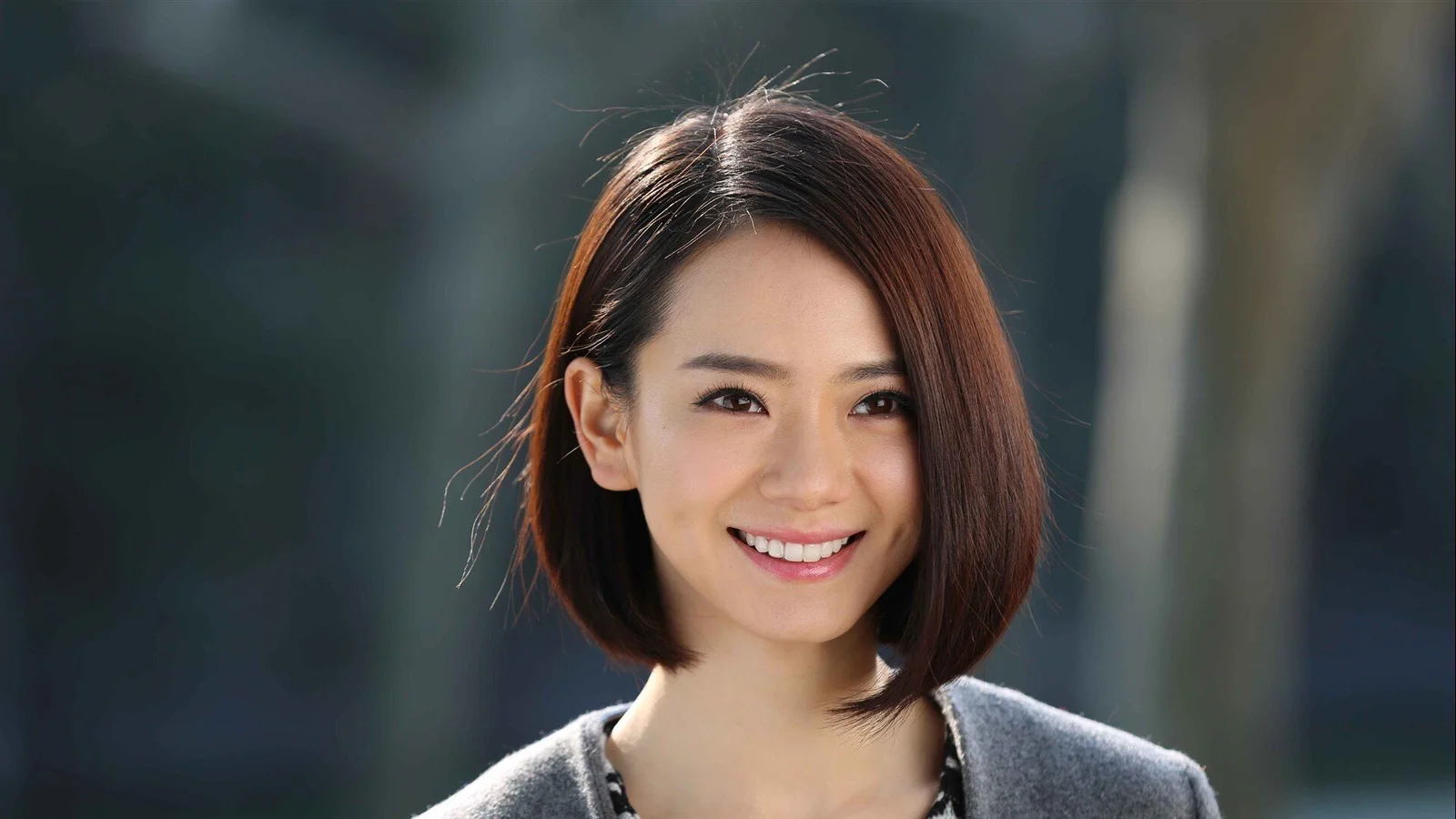 短い茶色の髪の女性が笑顔を見せ、灰色のトップスを着て、暗く柔らかくぼけた背景に立っています。