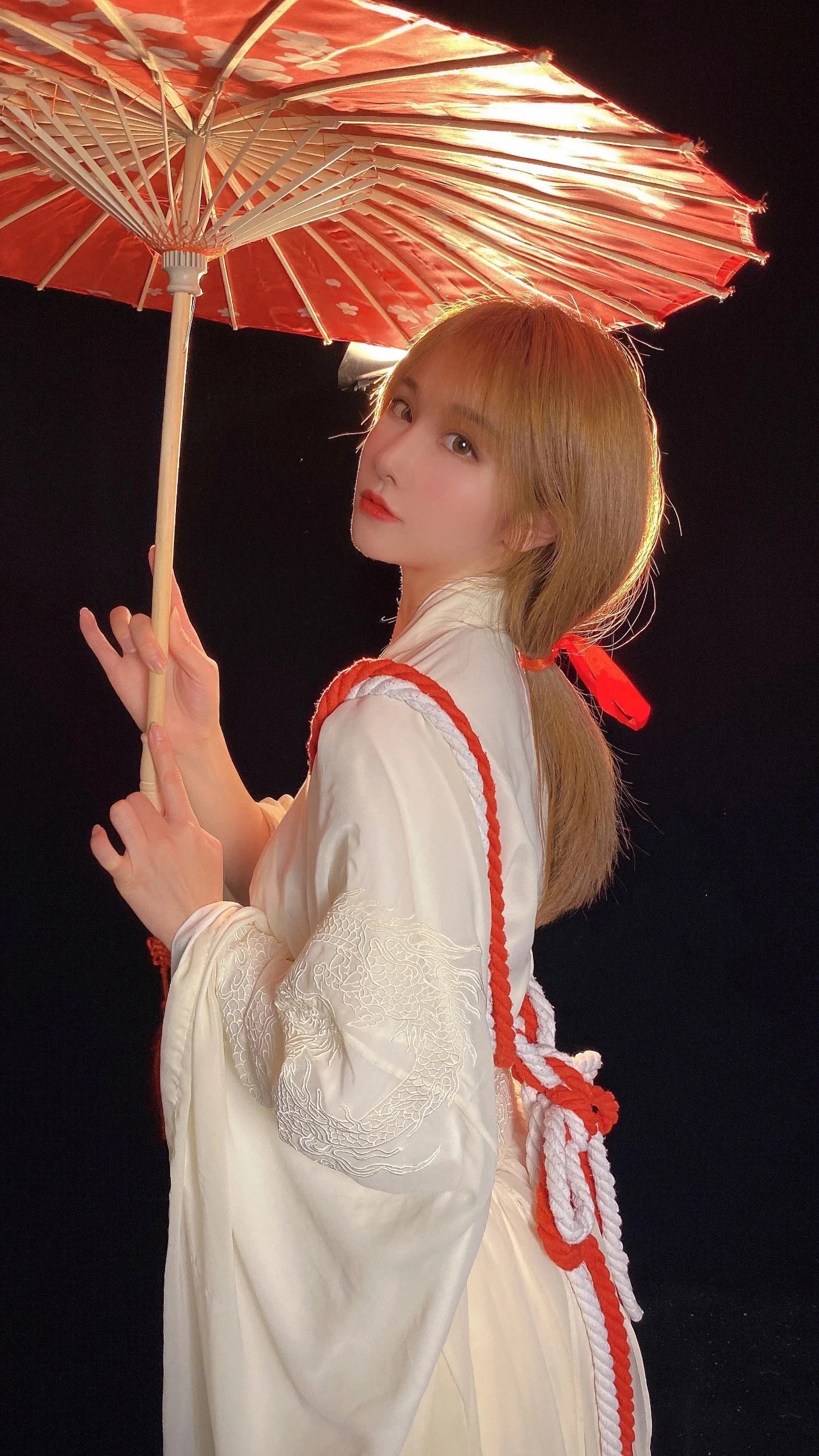 흰색 전통풍 의상을 입은 여성이 검은 배경 앞에서 붉은 양산을 들고 있습니다.