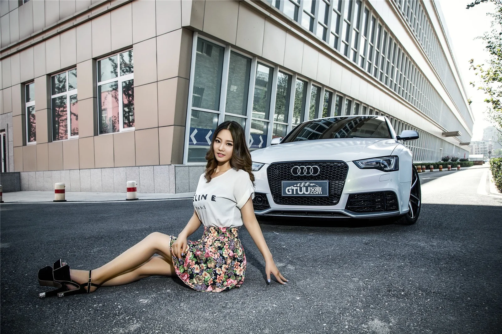一名成年女性坐在城市建筑前的道路上，身后停着一辆白色汽车。