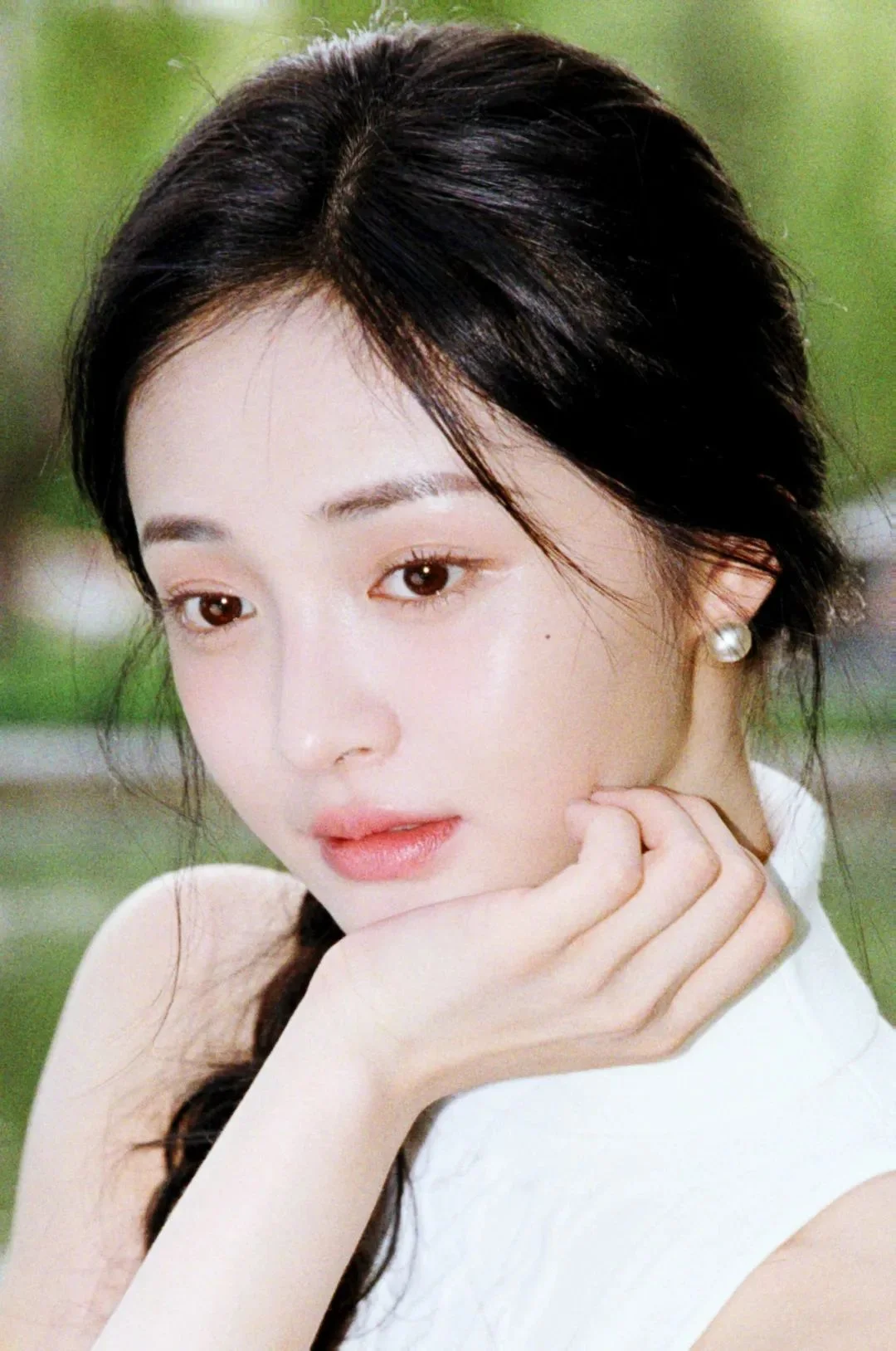 검은 머리 여성이 손으로 턱을 받치고 있는 클로즈업 인물 사진으로, 배경은 부드러운 녹색 흐림으로 보입니다.