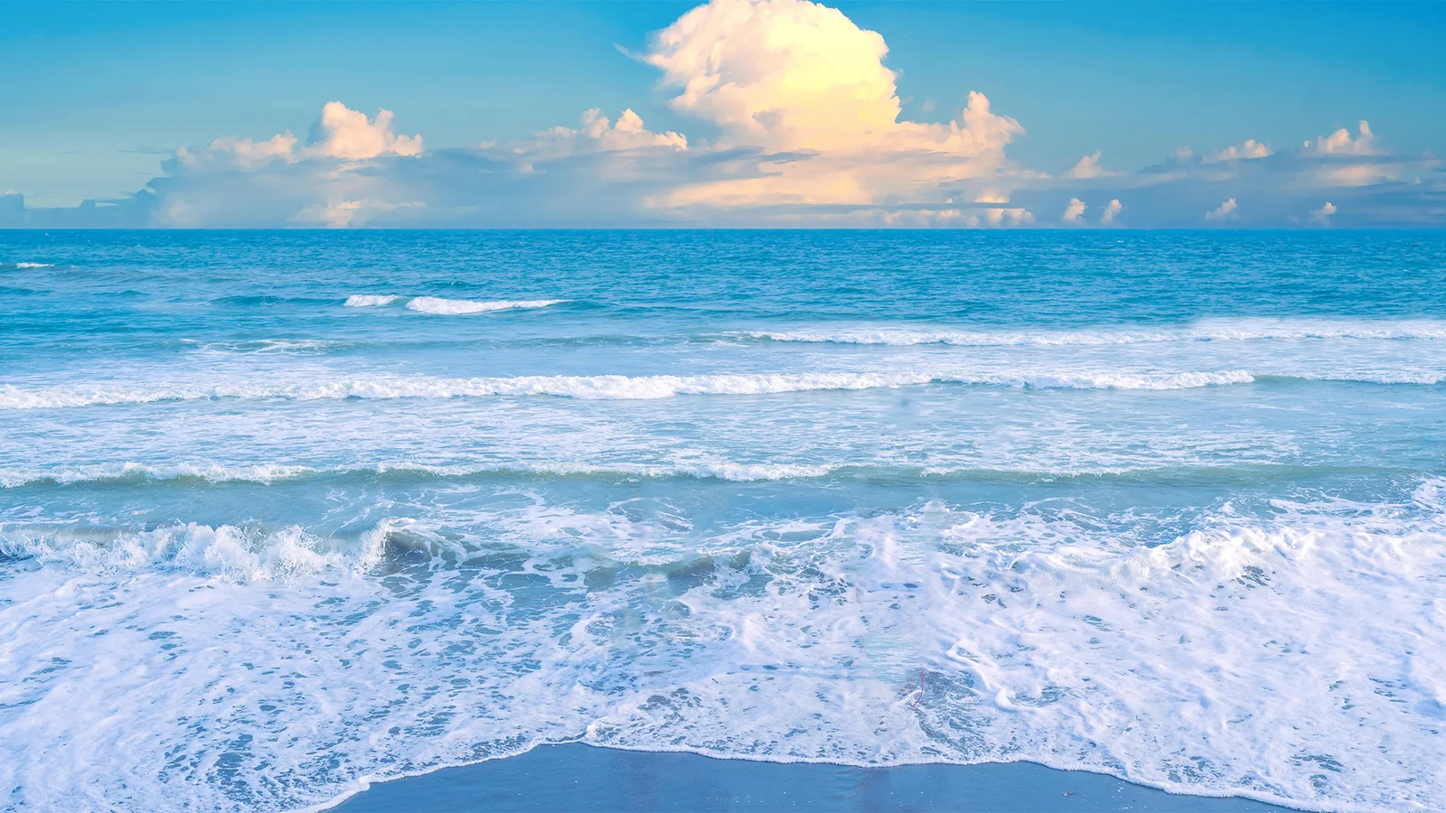 蓝色海面上层层海浪涌向沙滩，远处天空飘着白色云朵。