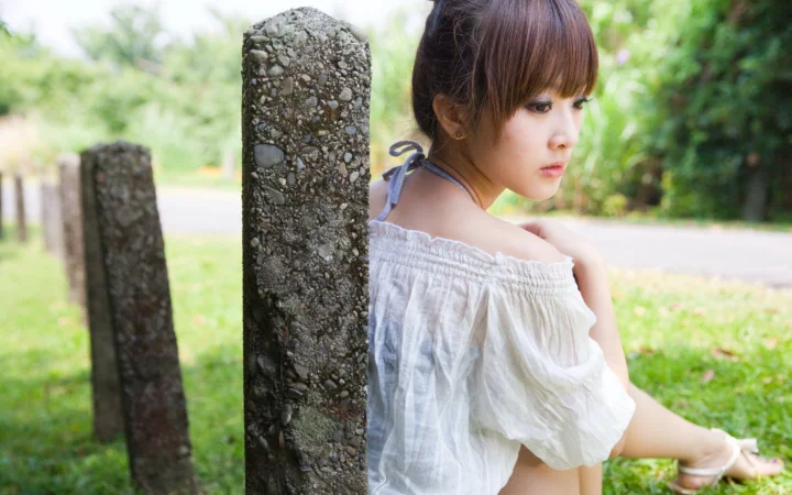 一名身穿白色上衣的女性坐在绿意环绕的户外石柱旁。