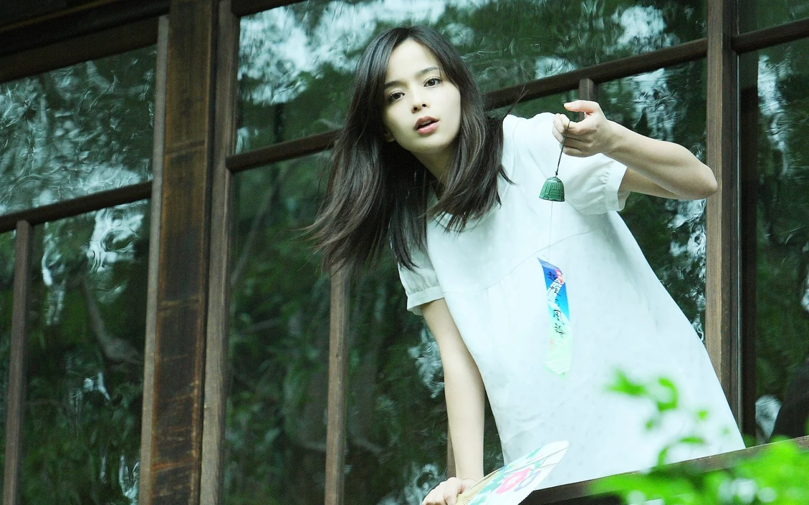 一名長髮女子穿著白色連衣裙站在窗邊，背景映著綠色植物。