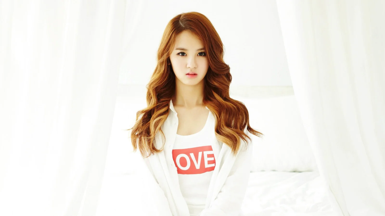 긴 머리의 여성이 문자가 있는 흰색 상의를 입고 밝은 흰색 실내 배경 앞에 앉아 있습니다.