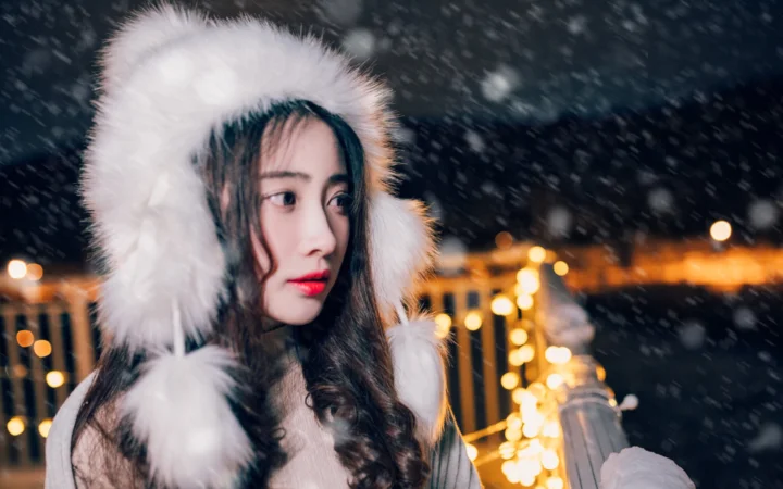 一名戴着白色毛绒帽的女子站在飘雪的夜晚，身后映着温暖的城市灯光。