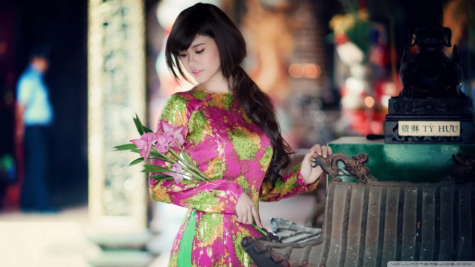 一名成年女性身穿粉綠色花卉傳統服飾，手捧鮮花站在室內雕像旁。