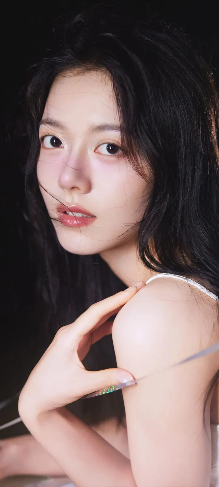 一位黑发女性在深色背景前侧身凝视镜头，手臂与肩部清晰可见。
