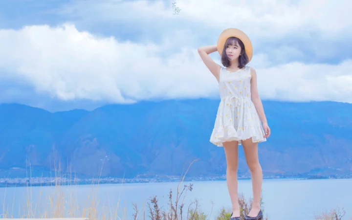 一名戴着草帽、穿白色连衣裙的女子站在湖畔，身后是蓝色山脉与天空。