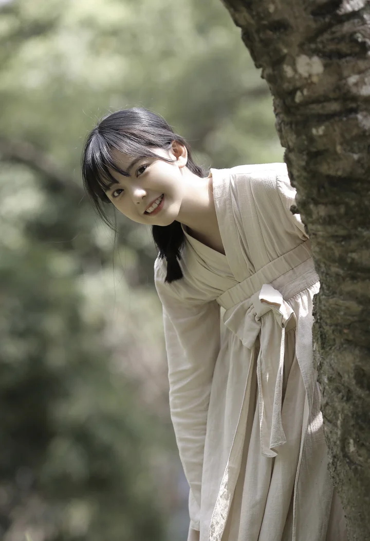 一名身穿浅色长裙的女子在森林中探身靠着树干微笑。