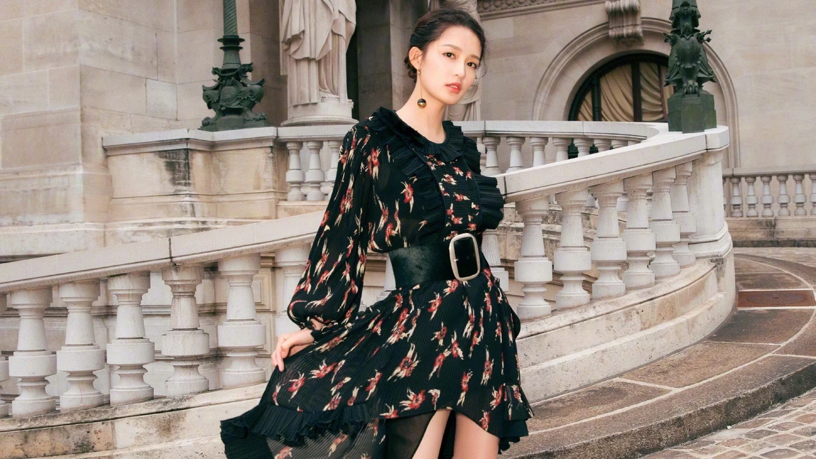 一名成年女性身穿黑色印花连衣裙，站在带有石 balustrade 的欧式建筑前。