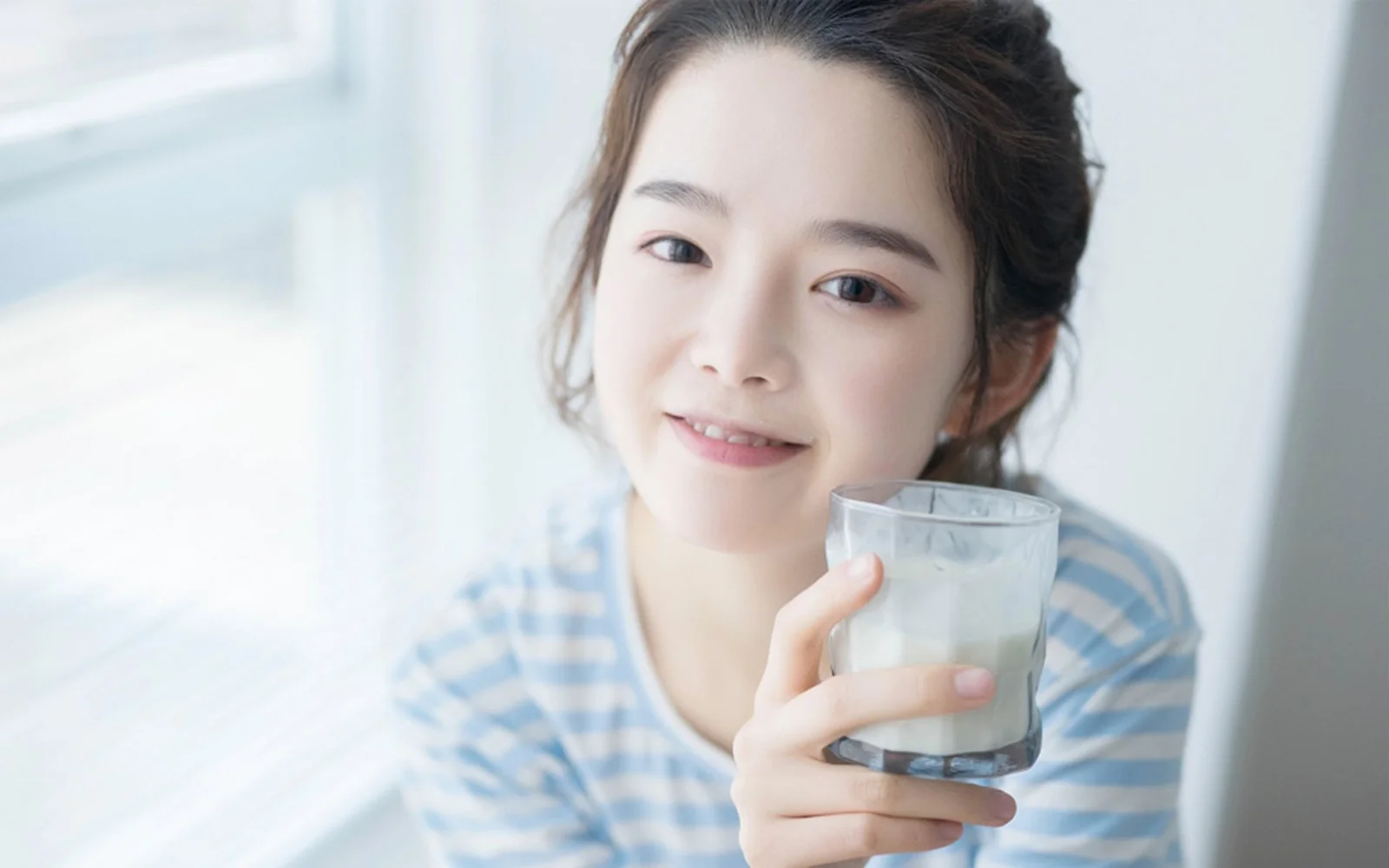 一名面带微笑的女子在明亮的窗边手持一杯牛奶。