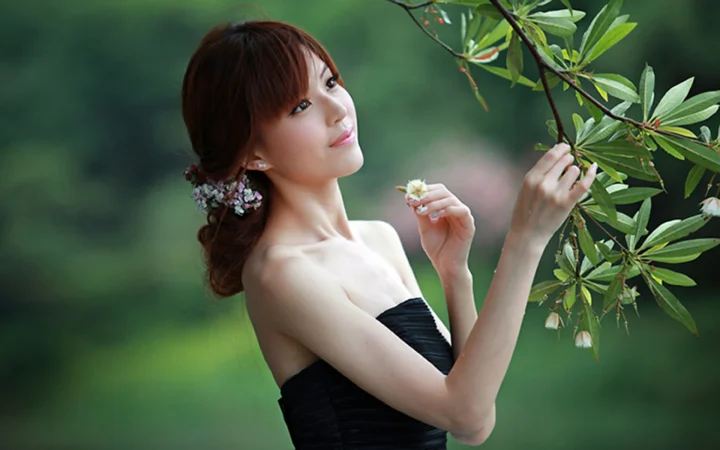 一名身穿黑色无肩带礼服的成年女性在绿叶树枝旁手持小花。