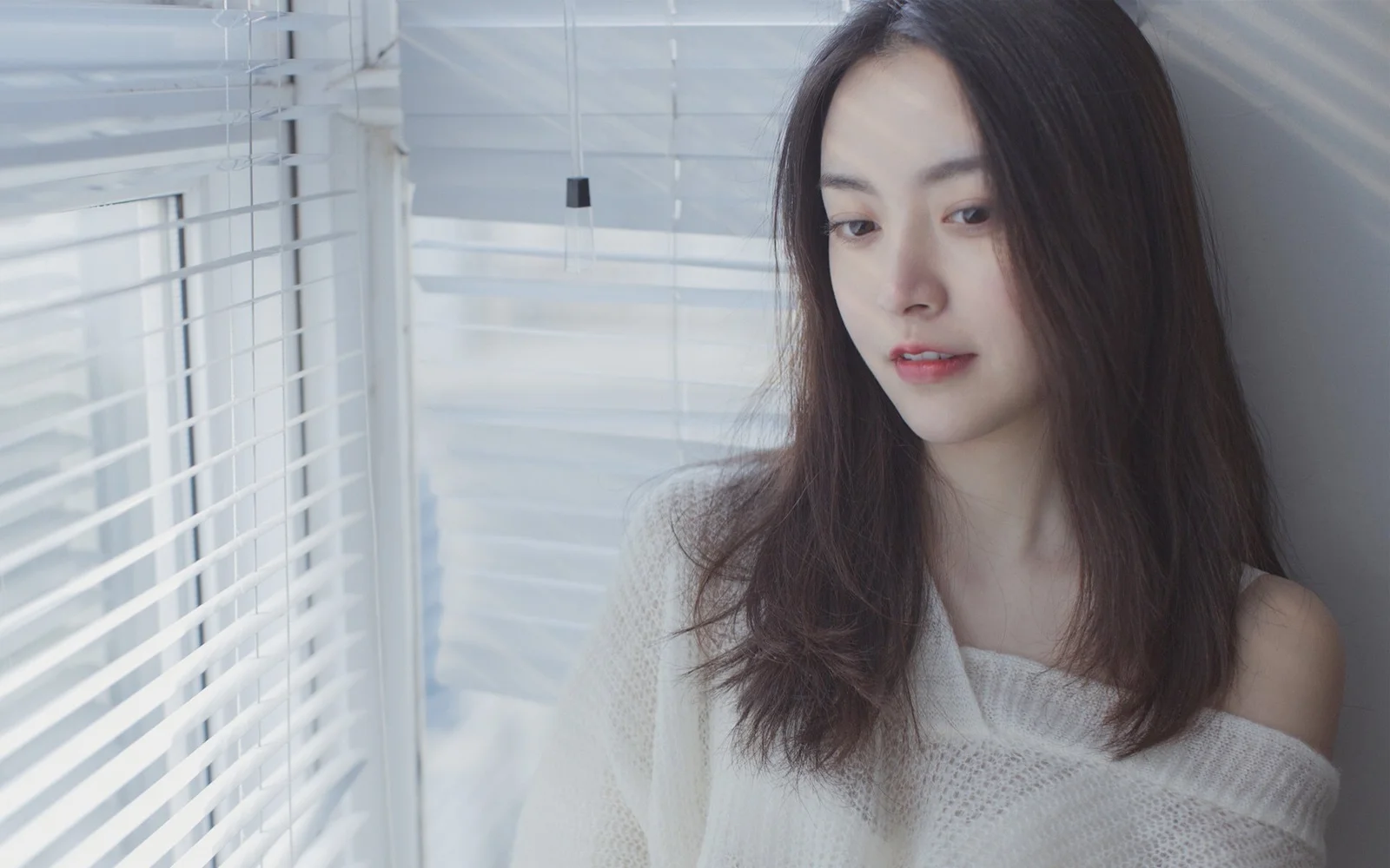 一名成年女性站在带百叶窗的窗边，柔和的自然光照亮了她的面容。