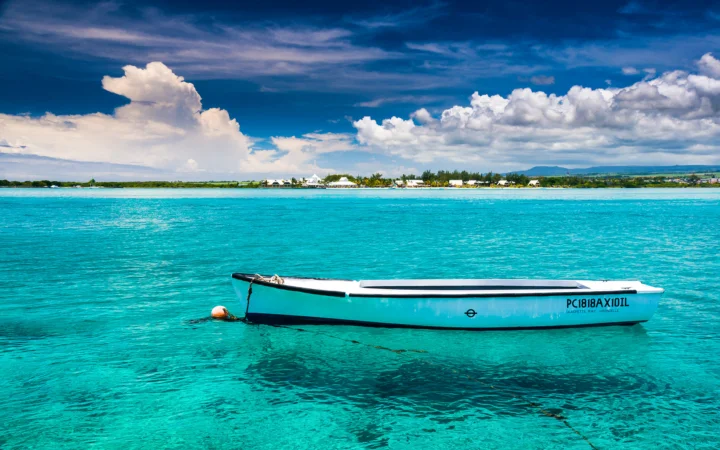 一艘浅蓝色小船漂浮在清澈的热带海水上，远处是海岸线和白云。
