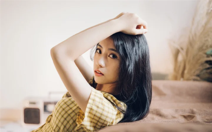 一名长发女子穿着格纹上衣坐在室内沙发旁，手臂抬起搭在头顶。