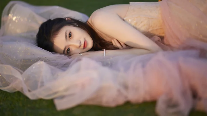 一名成年女性躺在草地上的浅色薄纱中注视镜头。