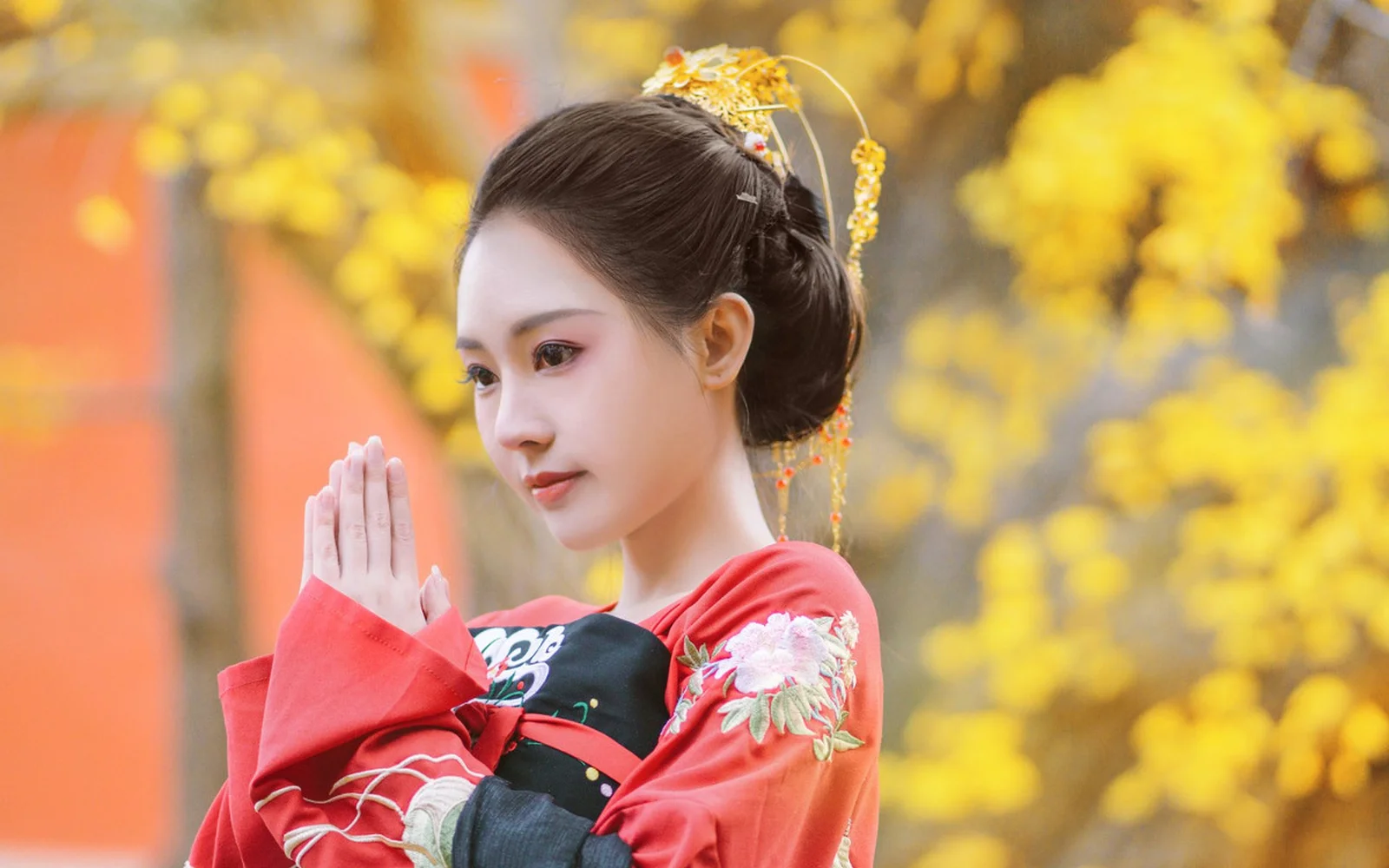 一名身着传统服饰的女子站在金黄色秋树前，双手合十。