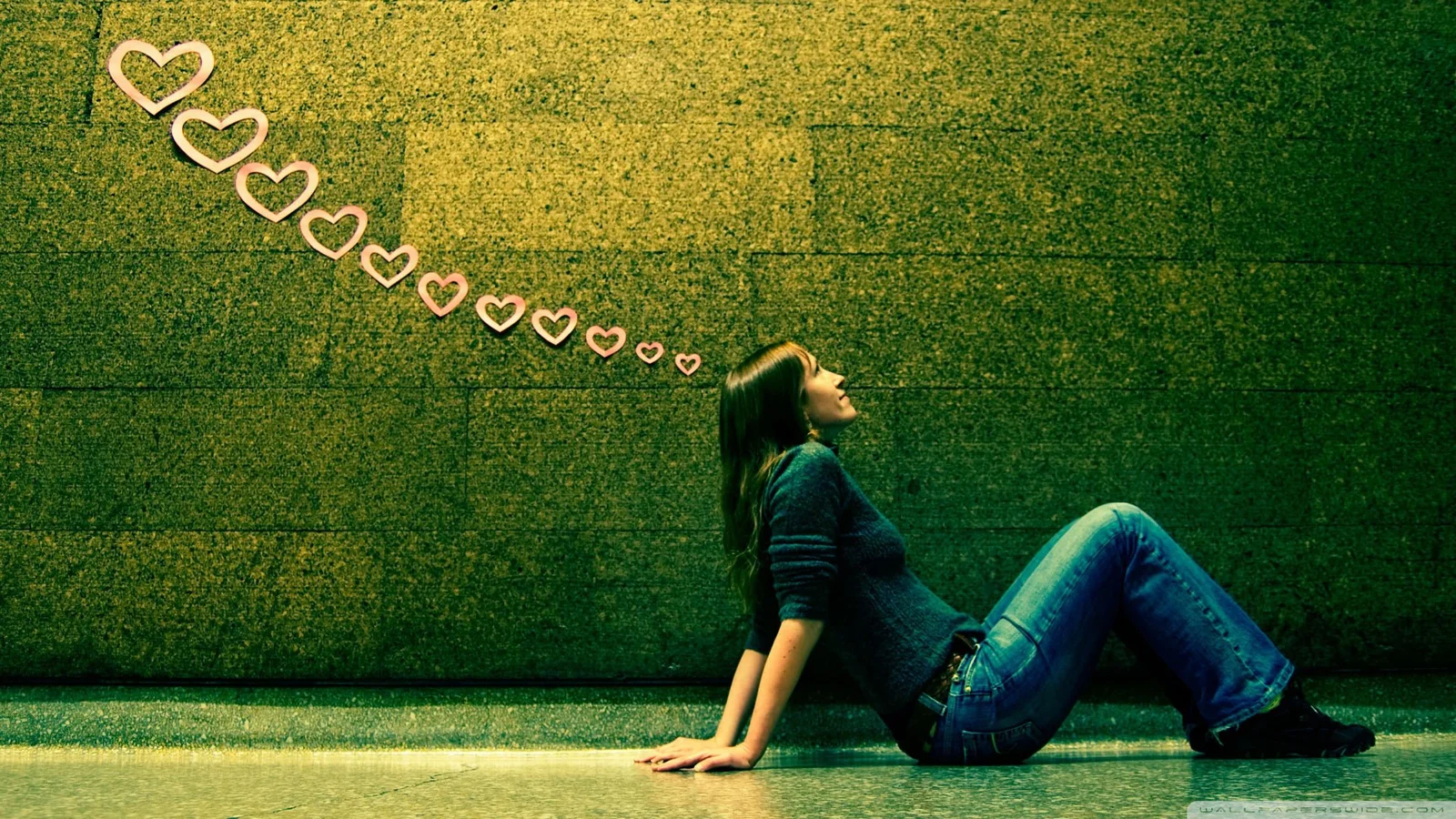 一名女子坐在深绿色墙面前，墙上方排列着一串心形图案。