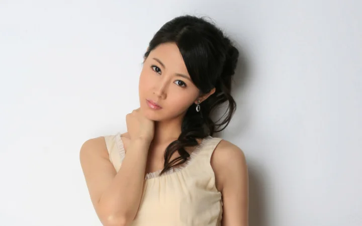 一名黑发女子穿着浅色无袖上衣靠墙站立，手托下巴望向镜头。