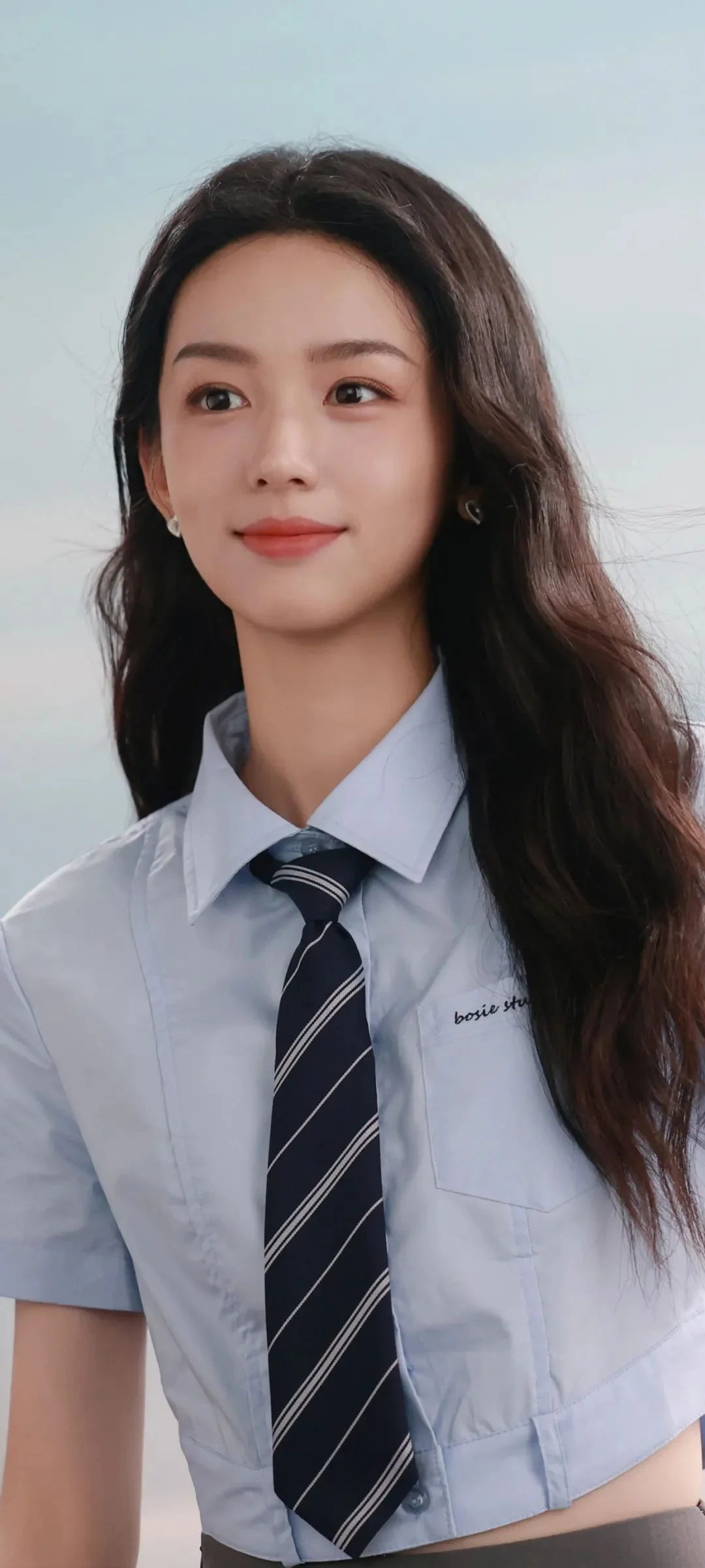 긴 머리의 여성이 밝은색 셔츠와 줄무늬 넥타이를 착용하고 카메라를 바라보며 미소 짓고 있다.