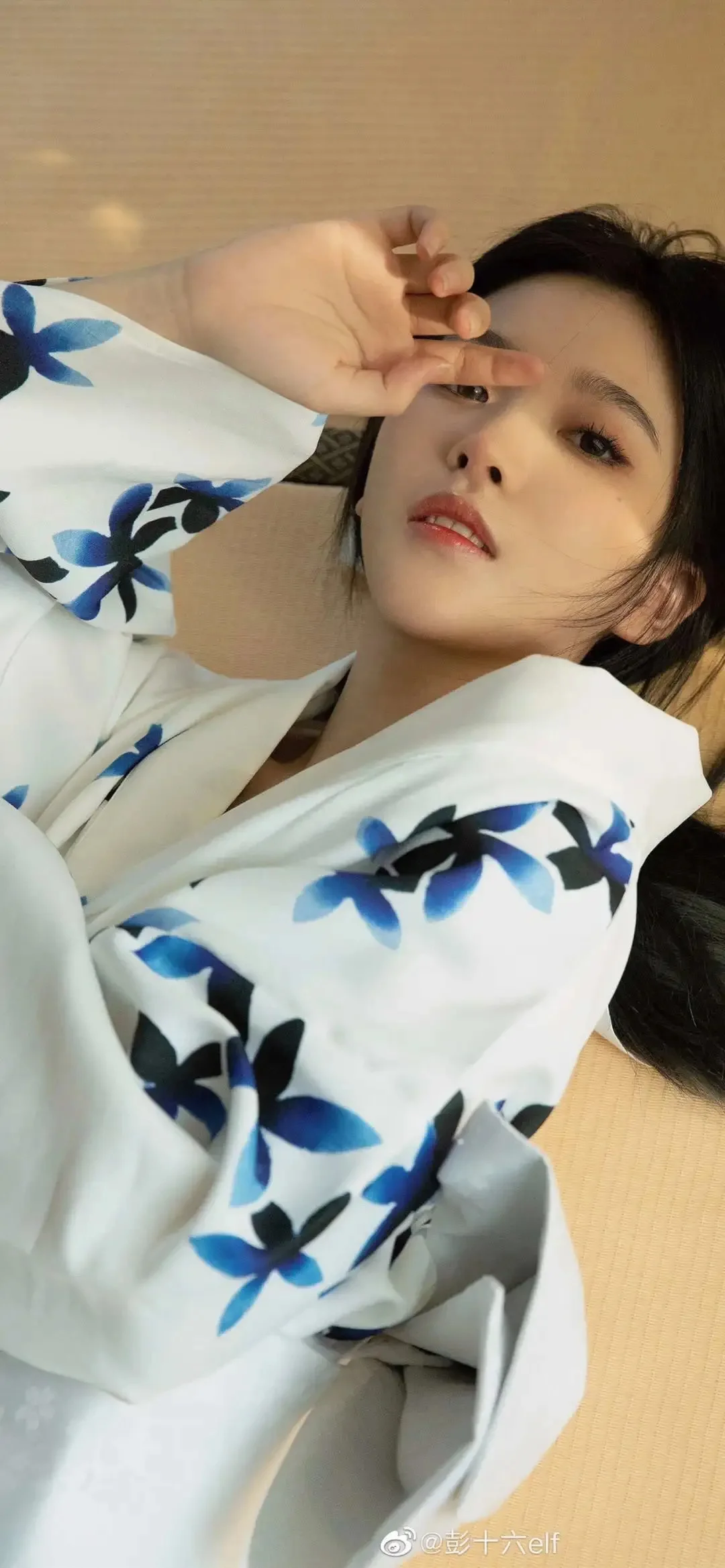파란색과 흰색 꽃무늬 옷을 입은 여성이 실내에 누워 한 손을 얼굴 가까이 들고 카메라를 바라보고 있다.